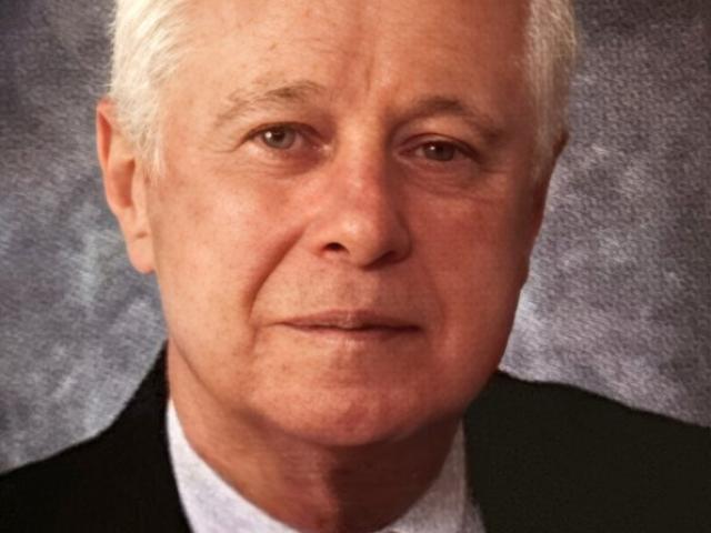 An older man with short white hair, wearing a black suit, white shirt, and red-and-black striped tie, poses against a textured gray background.