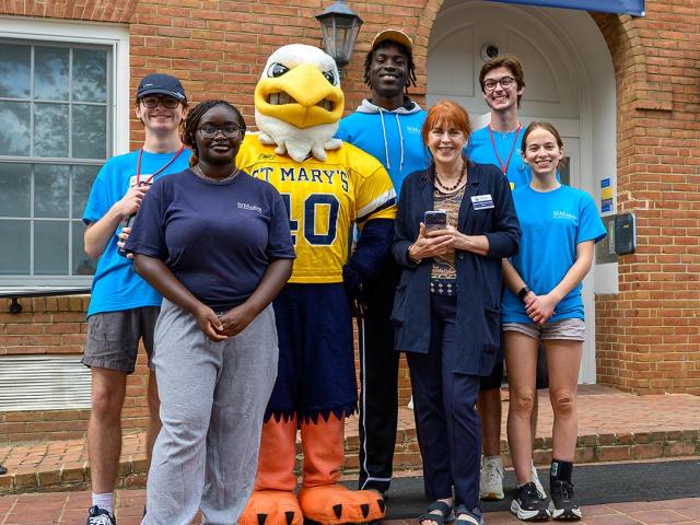 A group of six people and a person in a St. Mary's mascot costume pose in front of Dorchester Hall, smiling for the photo.