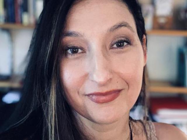 A woman with long brown hair and light makeup smiles softly at the camera, with bookshelves in the blurred background.