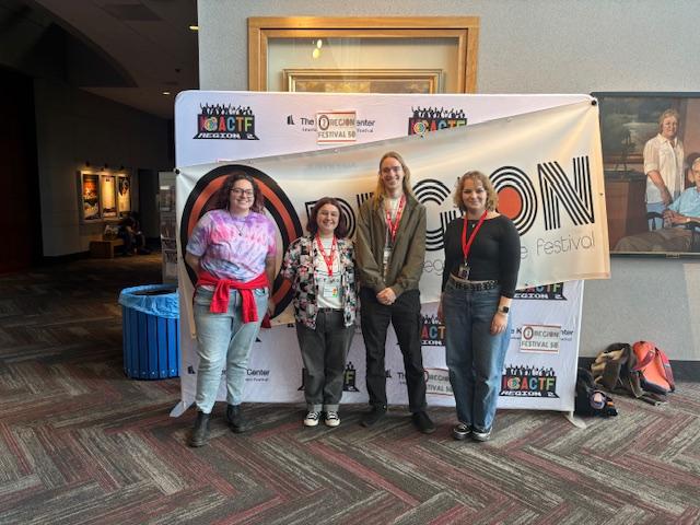 Four people with red lanyards stand in front of a banner for the American CollegeTheater Festival inside a building with framed art on the walls.
