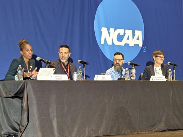 Four panelists sit at a table on stage with microphones, speaking in front of a blue backdrop displaying the NCAA logo.