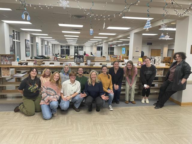 A group of students and a professor pose and smile for a photo in the Hilda C. Landers Library, with artist books and winter-themed decorations visible in the background.