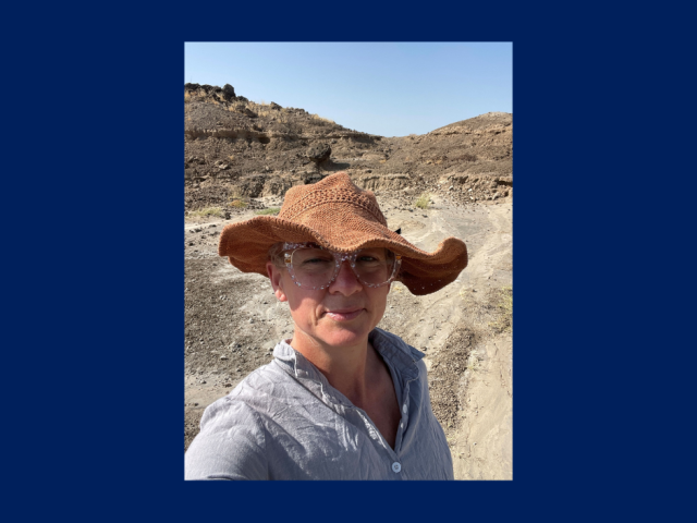 A person wearing a wide-brimmed hat, glasses, and a light shirt stands outdoors on a dusty path with rocky terrain in the background under a clear sky.
