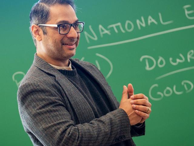 A man in glasses and a blazer stands in front of a green chalkboard with handwritten notes, gesturing as if speaking or teaching.