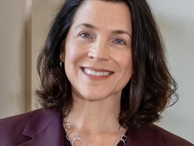A woman with shoulder-length dark hair, wearing a maroon blazer and a silver chain necklace, smiles at the camera against a neutral background.