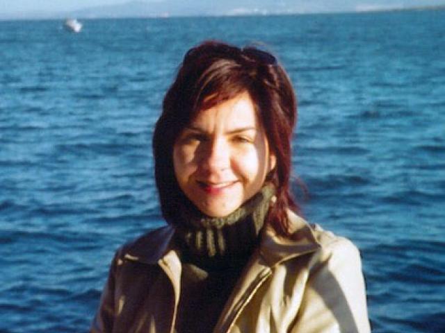 A woman with dark hair and a beige jacket stands in front of a large body of water with distant mountains in the background.