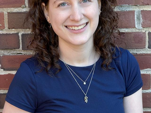 A woman with curly brown hair wearing a navy blue shirt and layered necklaces stands smiling in front of a brick wall.