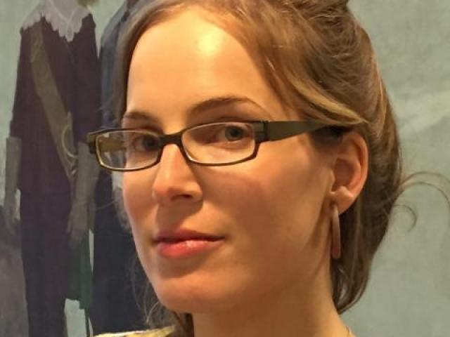 A woman with glasses and light brown hair pulled back looks at the camera. She is indoors, with a painting of two figures in historical dress in the background.