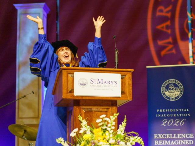 A person in academic regalia stands at a podium with arms raised, speaking at a St. Mary’s College of Maryland event, with a “Presidential Inauguration 2026” sign nearby.