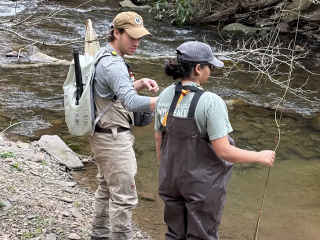 Two people in waders stand on a rocky riverbank with fishing gear, facing the water and preparing to fish in the stream.