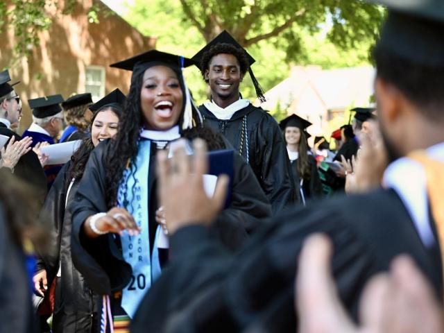 Graduates in caps and gowns walk in a procession outdoors, smiling and celebrating as people applaud around them.