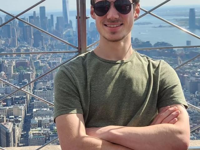 A man in sunglasses and a green shirt stands with arms crossed on an observation deck, with a city skyline and skyscrapers in the background.