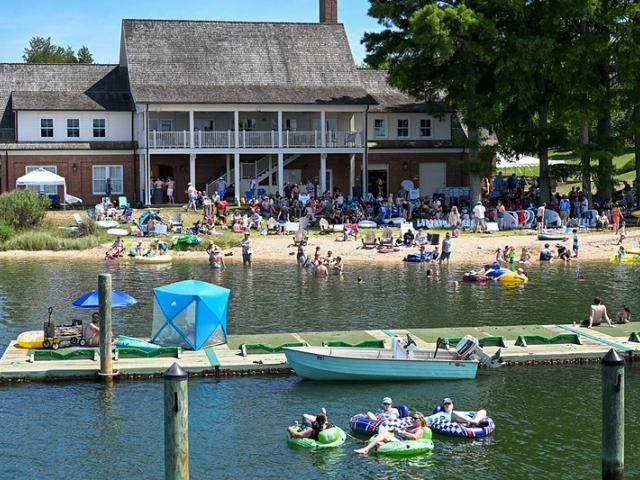 A crowd gathers at a lakeside beach near a large building, with people swimming, lounging on inflatables, and boats docked at a pier in the foreground.
