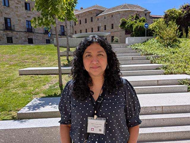 A woman with curly dark hair wearing a navy polka-dot blouse and name badge stands outdoors in front of stone steps and historic buildings on a sunny day.