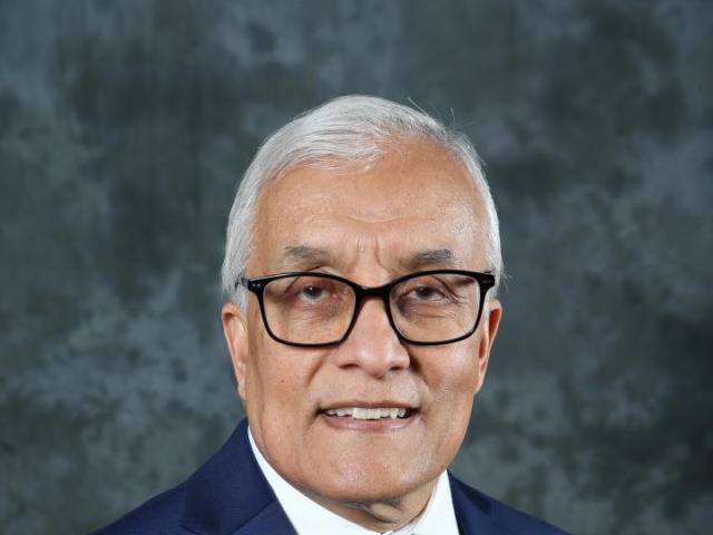 Older man with white hair and glasses, wearing a navy suit, white shirt, and green striped tie, posing in front of a mottled gray background.