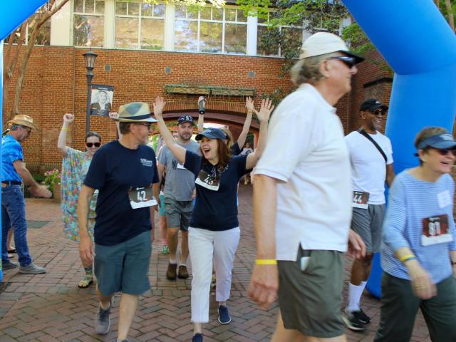 A group of people wearing race bibs walk under a blue inflatable arch outdoors; some are smiling and raising their hands in celebration.