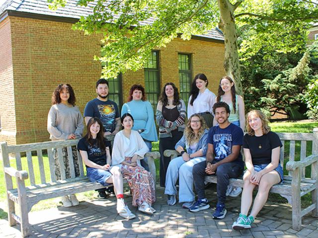 A group of eleven people pose for a photo outside on benches in front of a brick building and trees on a sunny day.