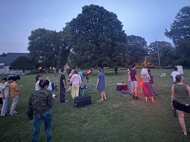 A group of people gathers on a grassy field at dusk, some standing near telescopes, while others form smaller groups. Trees and buildings are in the background.