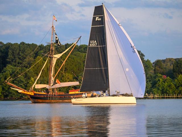 A sail boat with black and white sails passes the Maryland Dove talliship on the St. Mary's River 