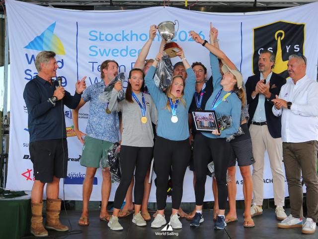 A group of people on stage celebrate with medals and a trophy at the World Sailing event, with banners and logos visible in the background. Photo by Malcom Hanes.