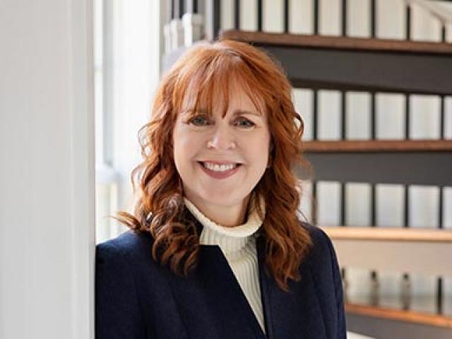 Woman with red hair wearing a navy blazer and white turtleneck stands smiling by a window, with shelves of binders in the background.