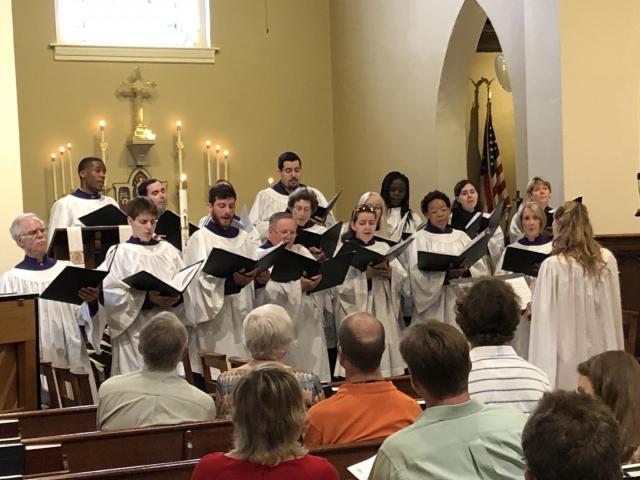 A choir robed in white singing at an old church altar in front of stained glass.