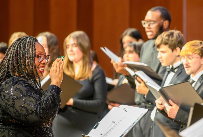 A choir conductor, wearing glasses and a black outfit, leads a group of choir singers.