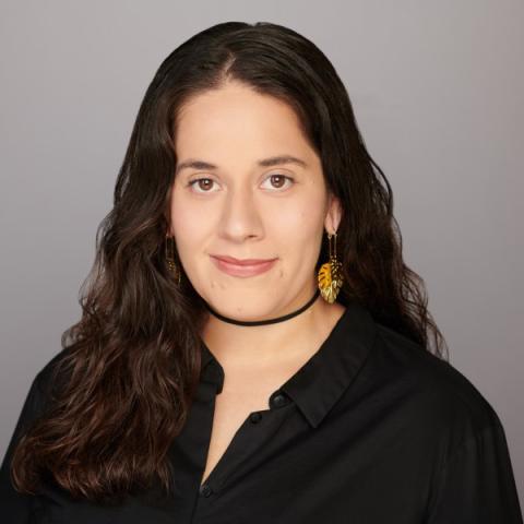 Person with long brown hair wearing a black collared shirt and leaf-shaped earrings against a plain background.
