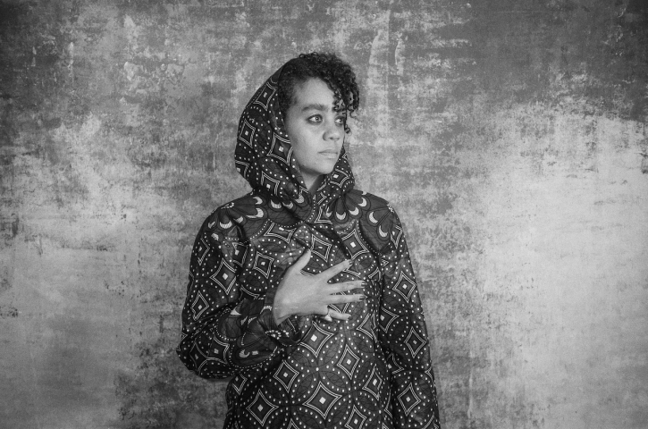 A person stands against a textured background, wearing a patterned hoodie. Their hand rests on their chest, and their eyes gaze thoughtfully into the distance. The image is in black and white.