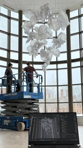 Two people stand on a scissor lift inside a glass-walled building, working on a large, abstract glass sculpture suspended from the ceiling. A laptop in the foreground displays a digital model of the sculpture.
