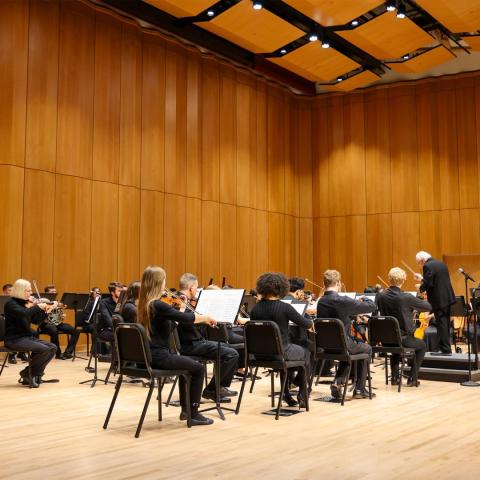 SMCM Orchestra performing on a concert stage with a conductor leading from the front in a wood-paneled hall.