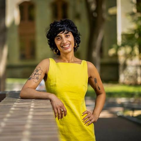 Person with short black hair in a bright yellow sleeveless dress, leaning on a brick wall outdoors. They have visible tattoos on both arms and are smiling, with a blurred background of trees and a building.