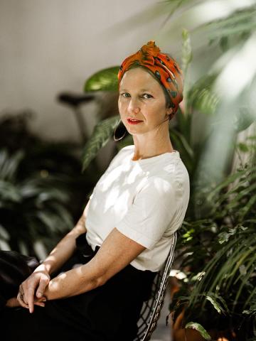 A woman sits amidst lush greenery, wearing a white T-shirt and a vibrant orange headscarf with patterns. She has hoop earrings and a thoughtful expression, bathed in soft sunlight filtering through the leaves.