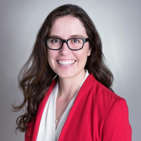 Woman with long brown hair wearing glasses, a red blazer, and a white blouse, smiling against a plain gray background.