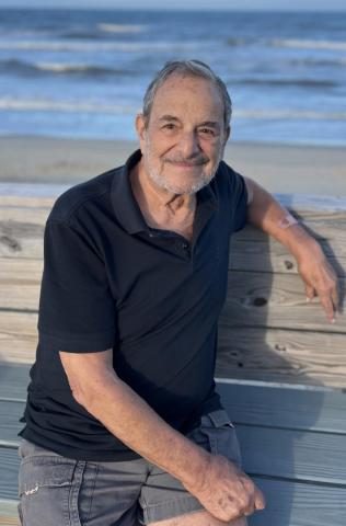 An older man with a beard and wearing a black polo shirt and shorts sits on a wooden bench by the beach, smiling at the camera.