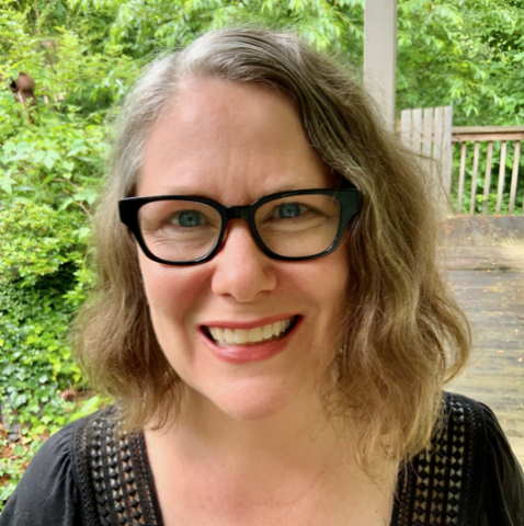 A person with glasses and shoulder-length wavy hair smiles outdoors, standing in front of greenery and a wooden deck.