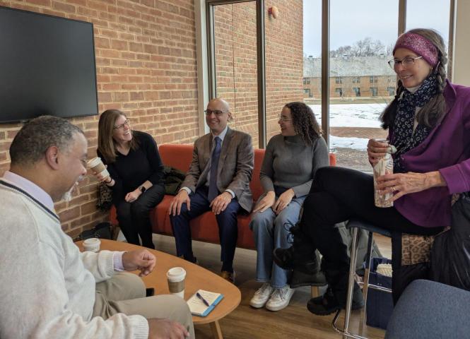 Five people seated and standing in a casual setting, talking and holding drinks. A snowy scene is visible through the window behind them.