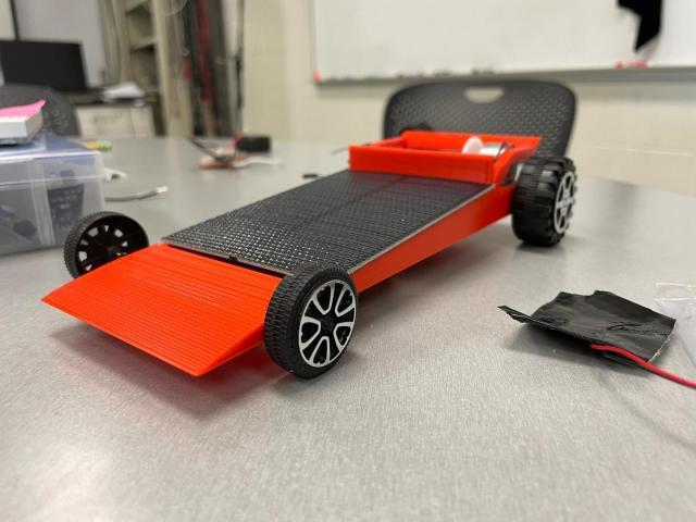A small, red model car with black wheels sits on a table in a classroom or workshop setting.