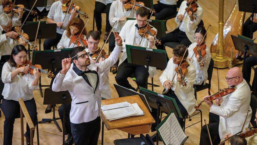 A conductor leads an orchestra of musicians playing string instruments. Most musicians are wearing white jackets and are focused on their sheet music.