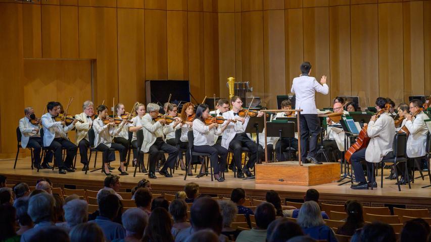 A conductor leads an orchestra of musicians in white jackets performing on a wooden stage in front of an audience.