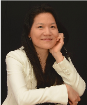A woman with long dark hair, wearing a white blazer, sits with her hand resting on her cheek and smiles slightly against a black background.