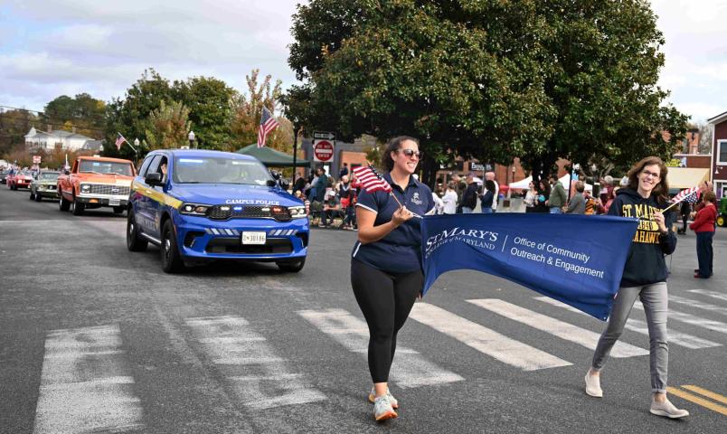People walk in a parade, holding a “St. Mary’s College of Maryland” banner, with a police vehicle and participants carrying American flags in the background.