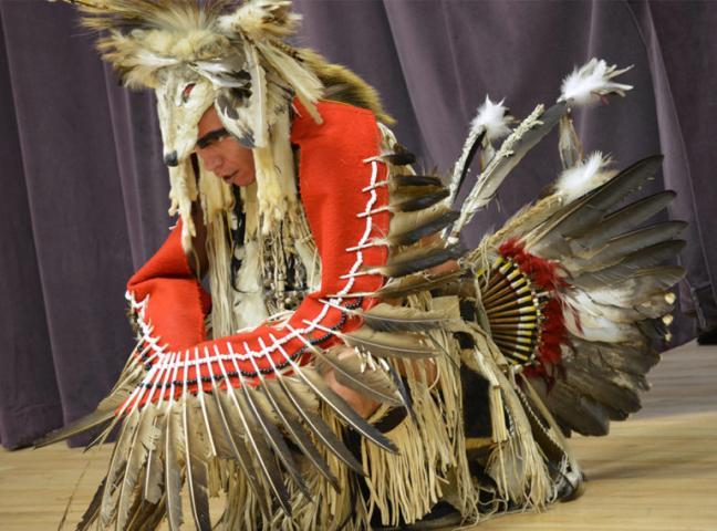 A person wearing traditional Native American regalia with feathers, animal fur, and a red garment kneels on a wooden floor in front of a purple curtain.