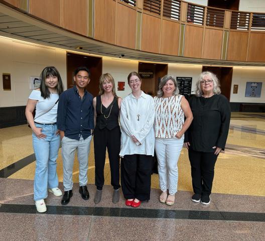 Six people stand side by side, posing for a group photo in a spacious, modern building with wood and marble accents.