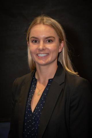 Claire Robey, a woman with straight blonde hair, wearing a dark blazer over a patterned blouse, smiles in front of a plain dark background.