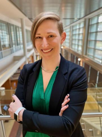 Rachel Romeo, with short hair in a black blazer and green top stands indoors with arms crossed, smiling in a modern, glass-walled building.