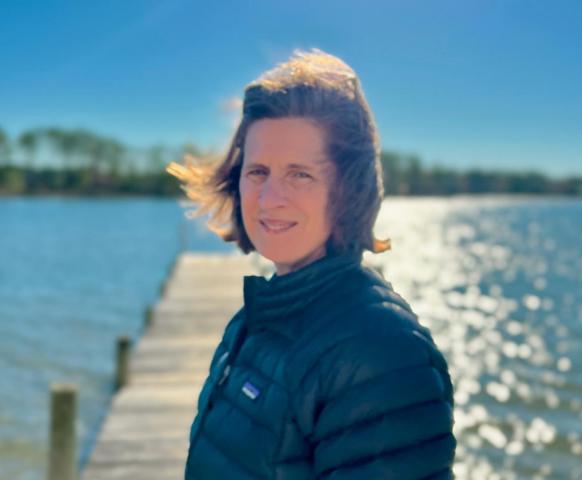 A person wearing a dark jacket stands on a wooden dock by a body of water, with sunlight reflecting off the water and trees in the background.