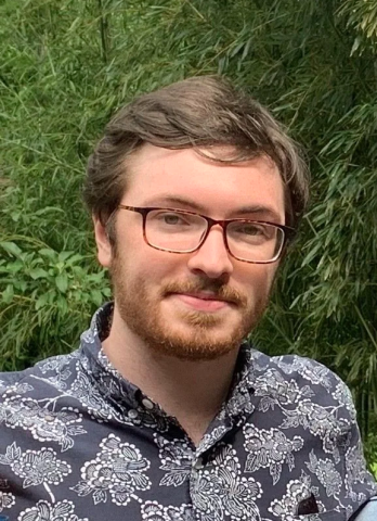 Max Madden, a man with glasses and a beard wearing a dark blue floral shirt stands outdoors in front of green foliage.