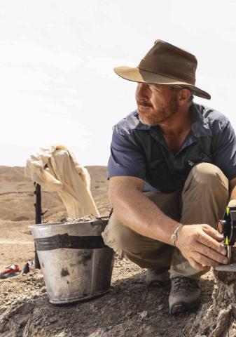 Man in a wide-brimmed hat squats outdoors next to a metal bucket, holding a tool, with rocky terrain and hills in the background.