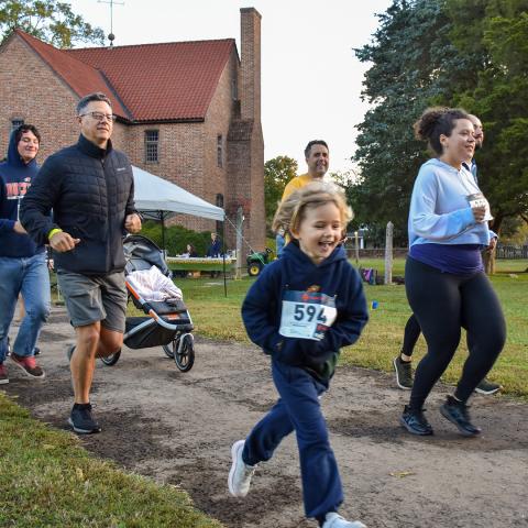 A group of people, including a young child with a race bib, jog together on a dirt path near a brick building during an outdoor race event.